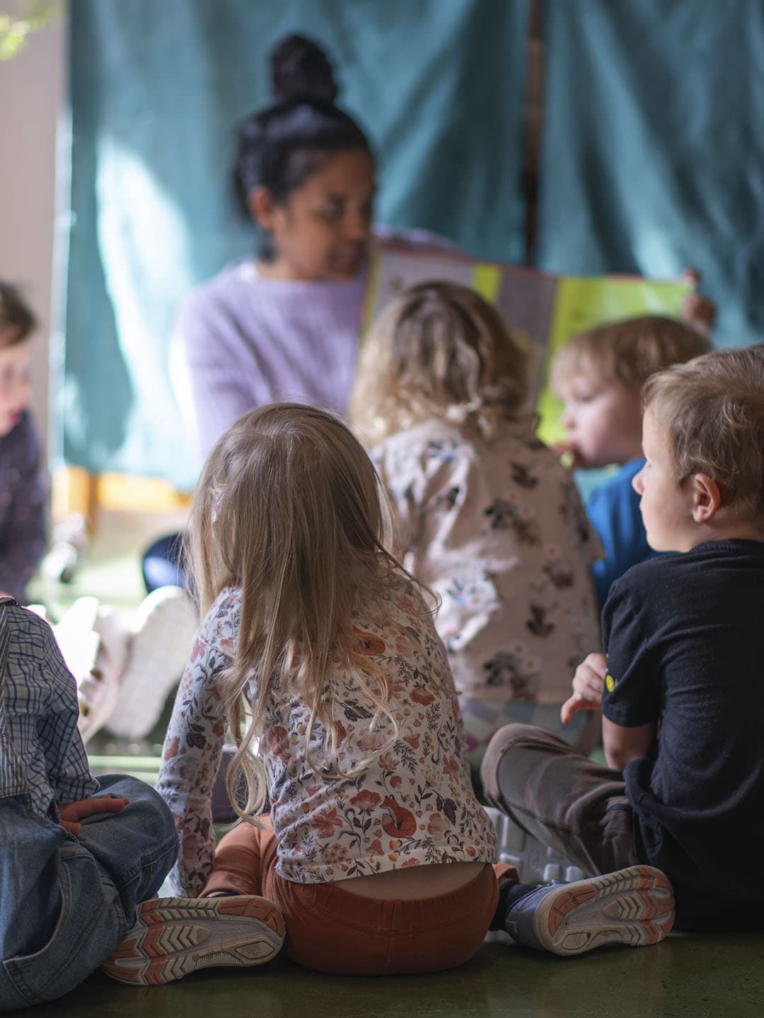 Children sitting together while a teacher reads in a daycare classroom.